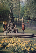 Load image into Gallery viewer, Children playing around a statue in a park with yellow flowers in the foreground.