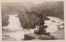 Load image into Gallery viewer, Person sitting on a rock by a flowing stream with a mountainous background