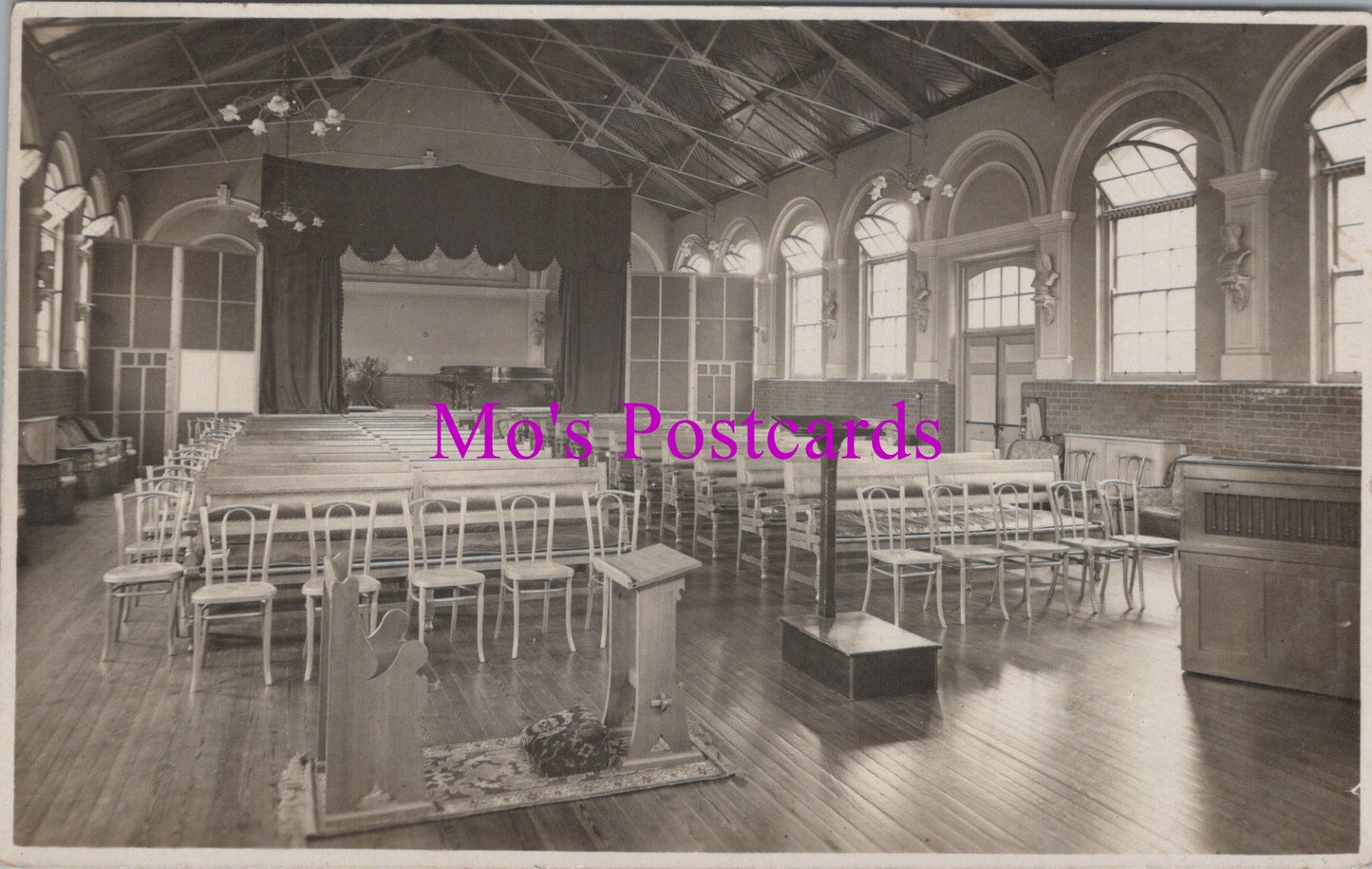 Vintage black and white photo of a large room with rows of chairs and a stage, labeled 'Mo's Postcards'.