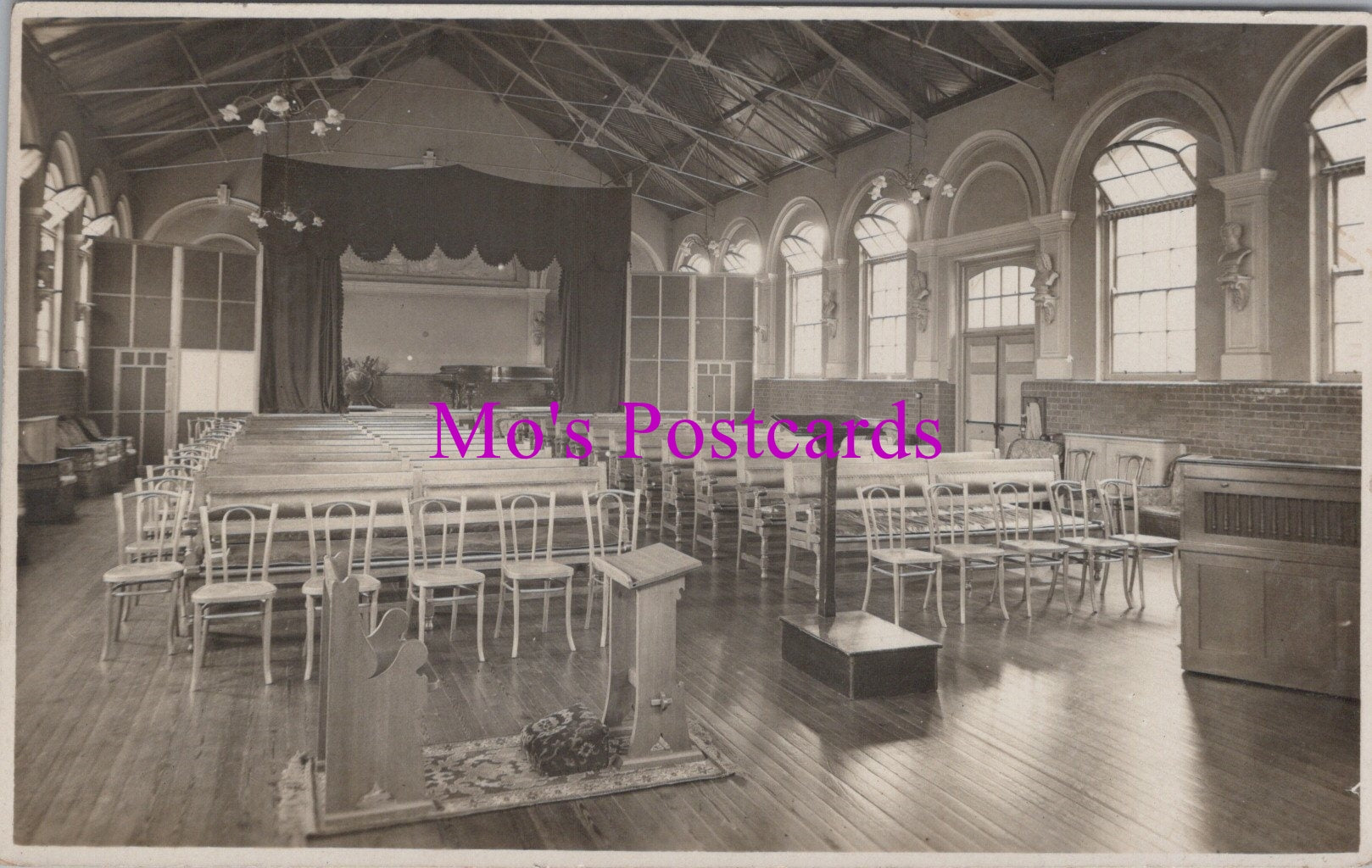 Vintage black and white photo of a large room with rows of chairs and a stage, labeled 'Mo's Postcards'.