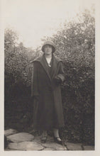 Load image into Gallery viewer, Person wearing a long coat and hat standing outdoors with greenery in the background
