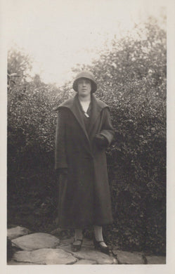 Person wearing a long coat and hat standing outdoors with greenery in the background