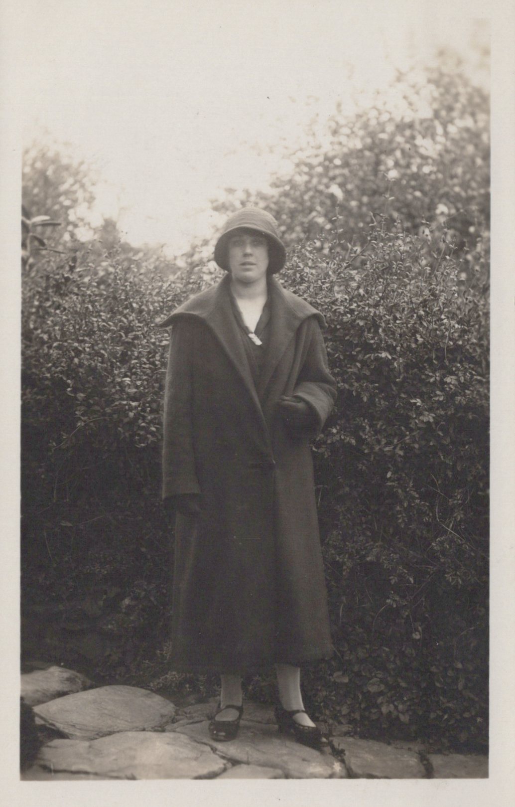 Person wearing a long coat and hat standing outdoors with greenery in the background