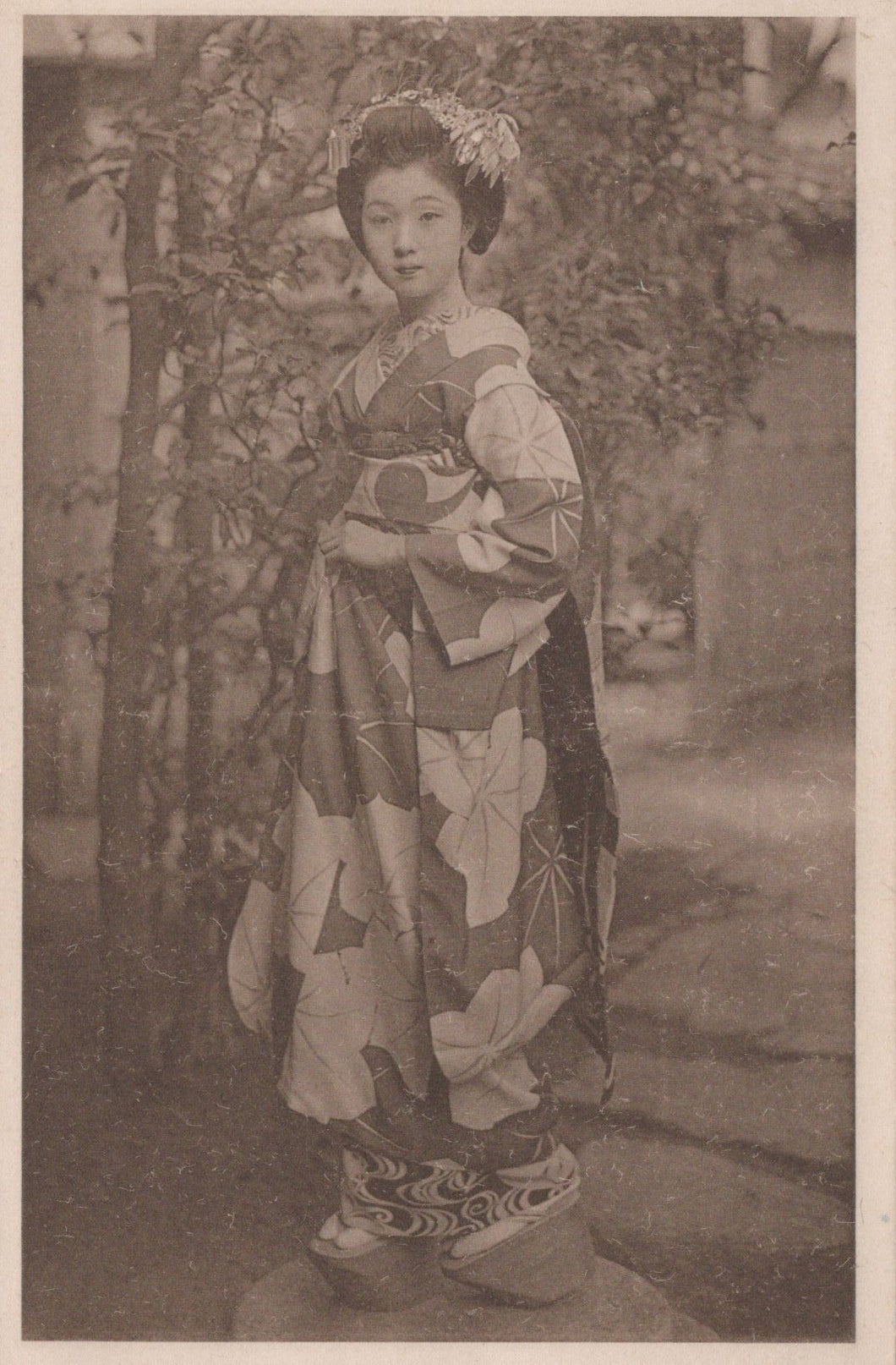 Woman in a traditional kimono standing outdoors with trees and flowers in the background