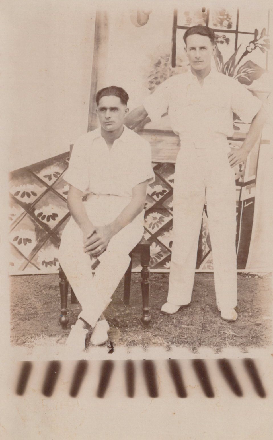 Two men in cricket uniforms standing outdoors with a decorative background