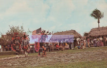 Load image into Gallery viewer, Traditional dancers with an American flag in a grassy area with huts and people in the background.
