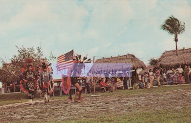 Traditional dancers with an American flag in a grassy area with huts and people in the background.