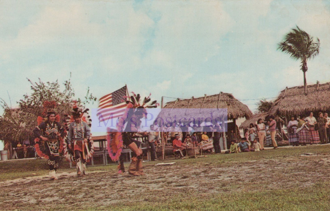 Traditional dancers with an American flag in a grassy area with huts and people in the background.