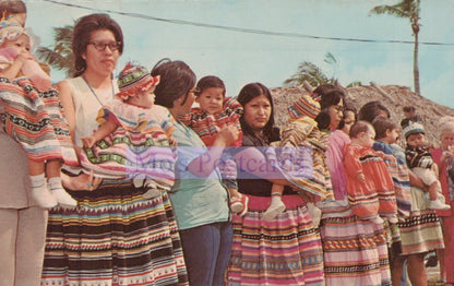 Group of women in traditional clothing with children on their backs, set against a tropical background.