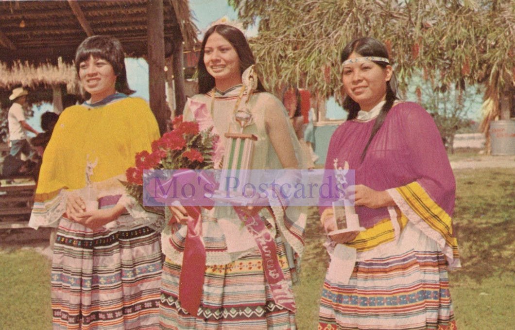 Three women in traditional attire standing outdoors with a 'Mo's Postcards' logo overlay.