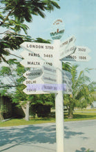 Load image into Gallery viewer, Directional signpost with distances to various cities in the background of a tropical setting.

