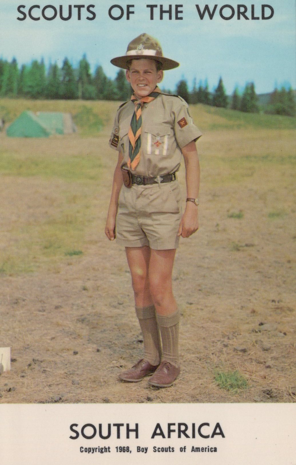 Scout in uniform standing outdoors with trees and tents in the background, labeled 'Scouts of the World, South Africa'.