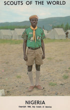 Load image into Gallery viewer, Scout in Nigeria wearing a green uniform with a yellow scarf, standing in an open area with buildings in the background.
