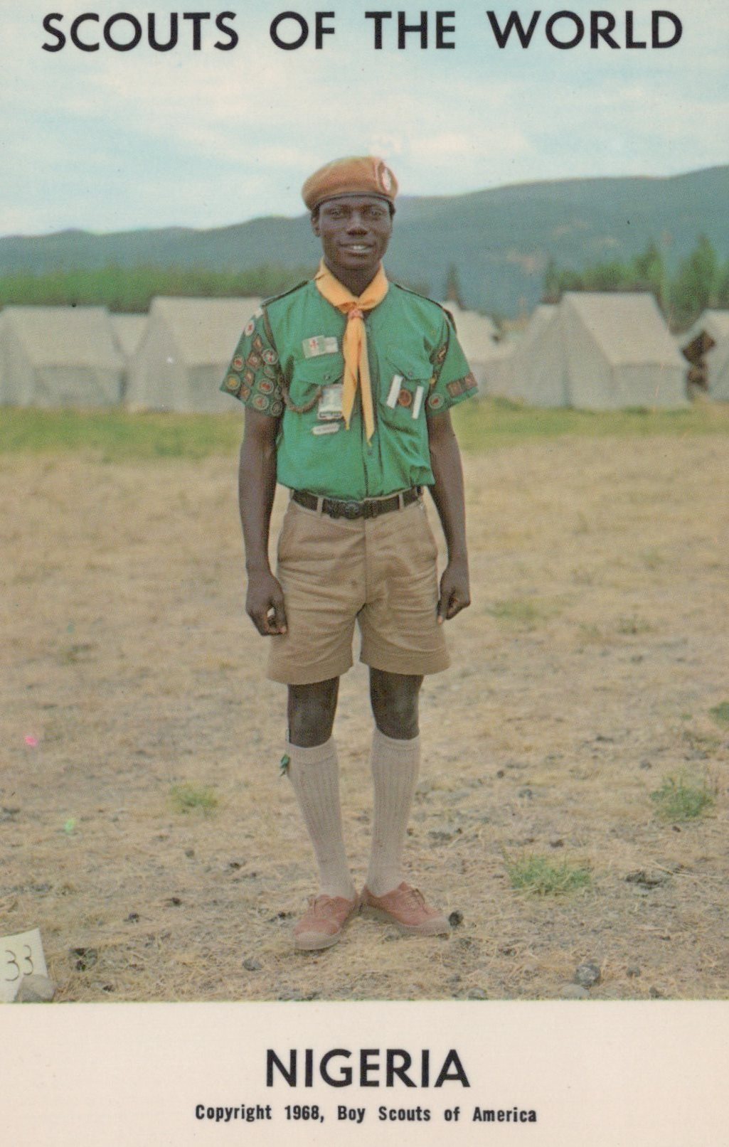 Scout in Nigeria wearing a green uniform with a yellow scarf, standing in an open area with buildings in the background.