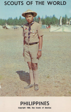 Scout in uniform with a badge, standing outdoors with trees and tents in the background.
