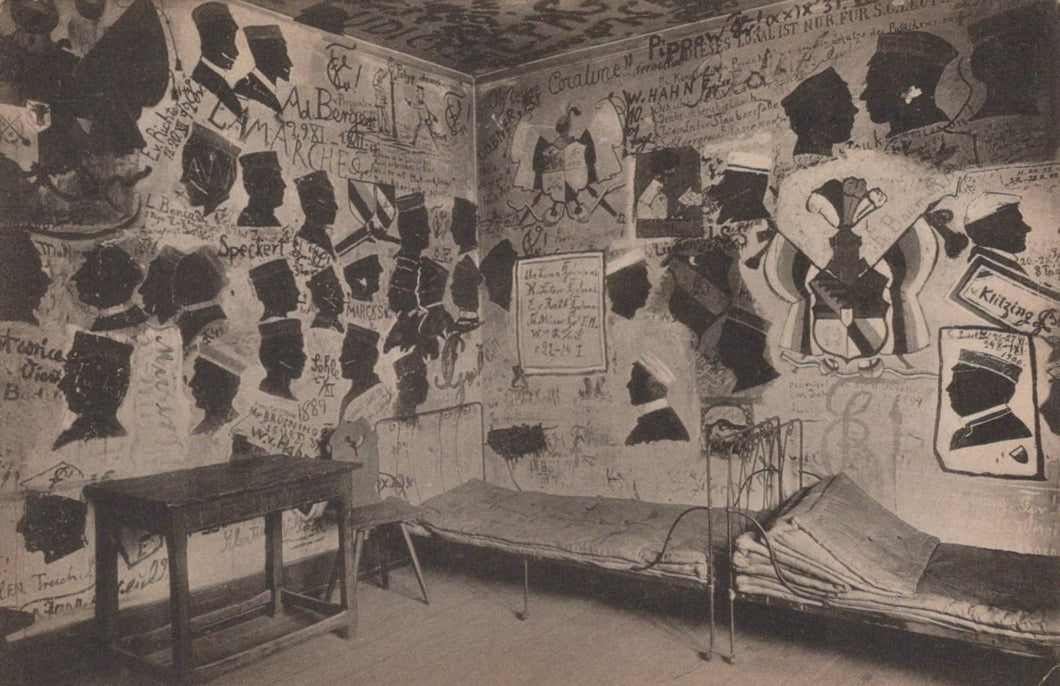 Room with walls covered in posters and notices, featuring a bed and table.