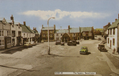 Vintage photograph of a town square with buildings and parked cars under a blue sky.