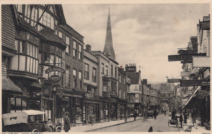 Vintage black and white photo of a street scene with shops and a church spire.