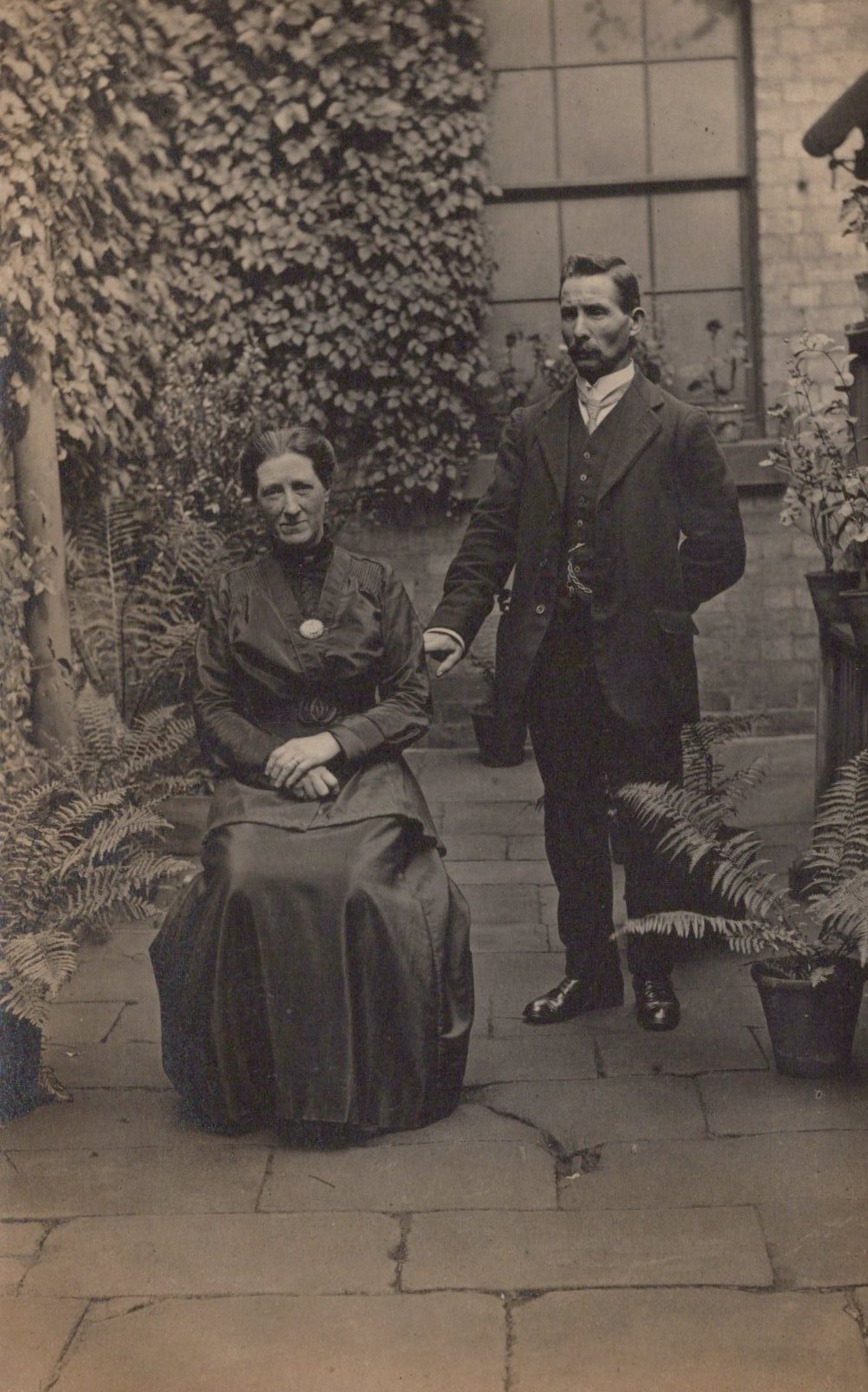 Vintage black and white photograph of a man and woman outdoors with plants and a building in the background.