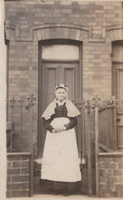 Load image into Gallery viewer, Woman in a white apron standing in front of a door with a brick wall background
