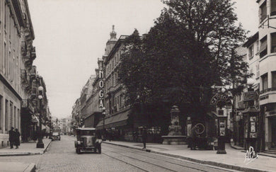 Vintage black and white photograph of a city street with buildings, a car, and pedestrians.