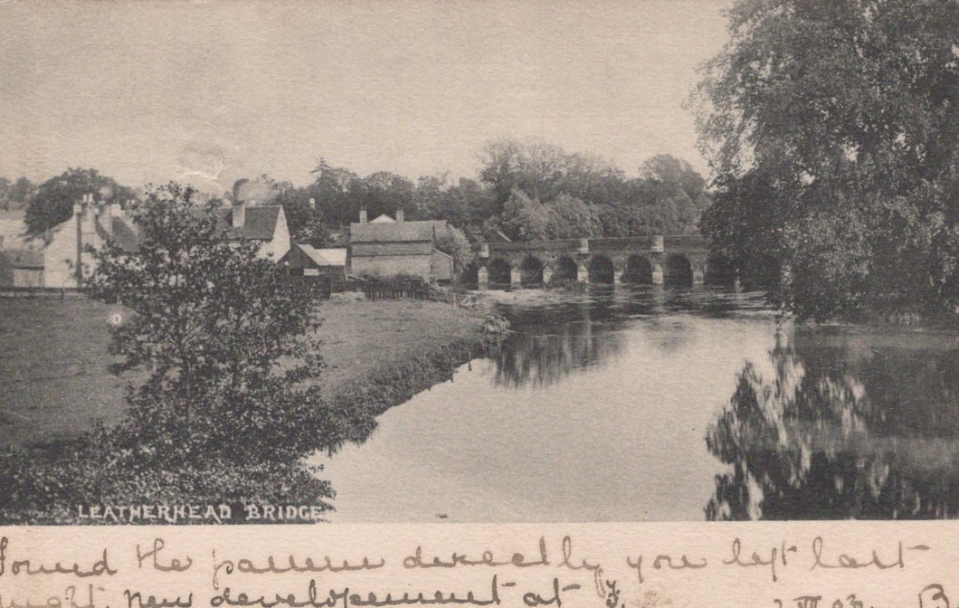 Vintage black and white photograph of a bridge over a river with trees and buildings in the background.