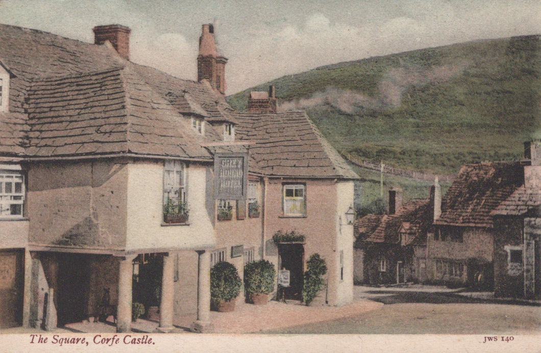 Vintage postcard of a village square with houses and a sign, featuring a steam train in the background.
