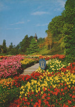 Load image into Gallery viewer, Colorful flower garden with a pathway leading to a scenic view of trees and a tower.
