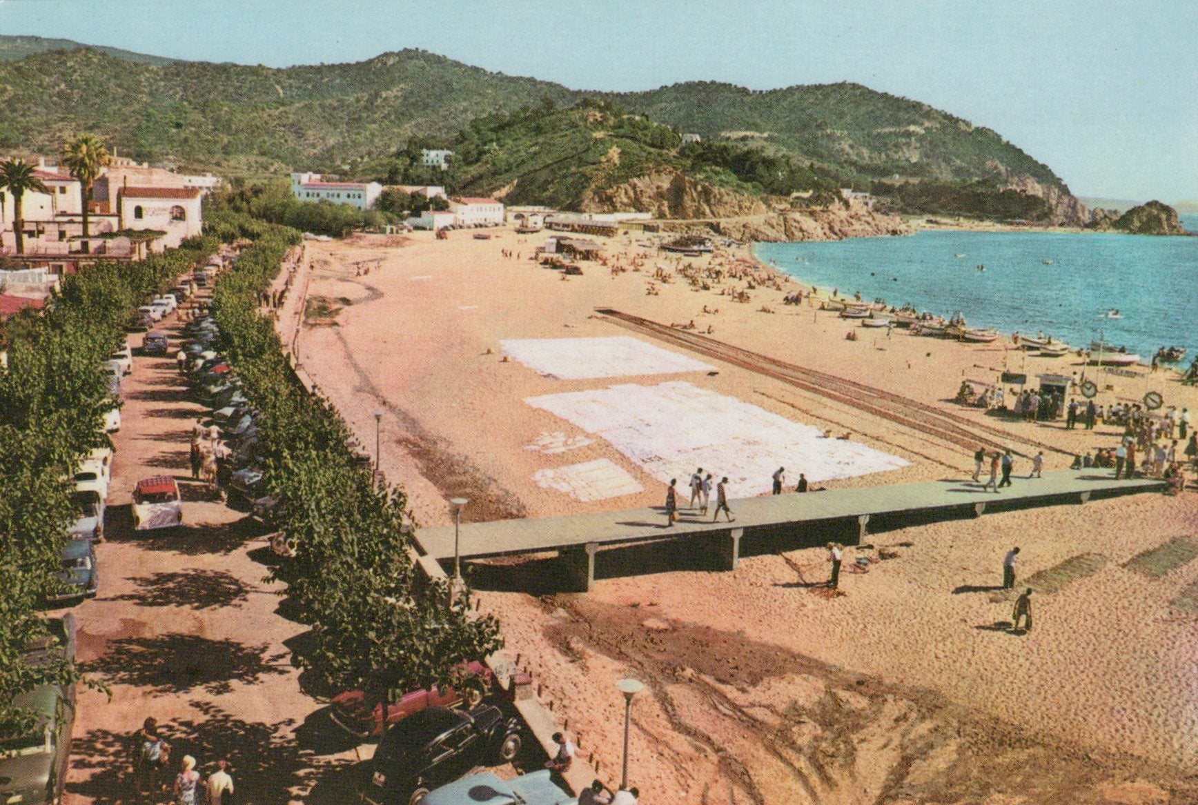 Beach scene with people, cars, and buildings along a road next to a sandy beach.