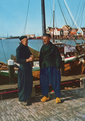 Two men standing on a dock with boats and a village in the background
