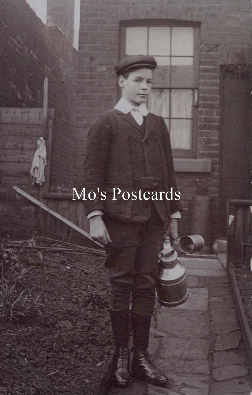 Vintage black and white photo of a boy holding a water jug in front of a brick building.