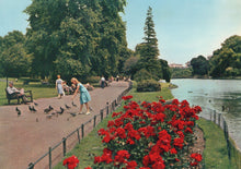 Load image into Gallery viewer, People walking along a path by a lake with red flowers in the foreground
