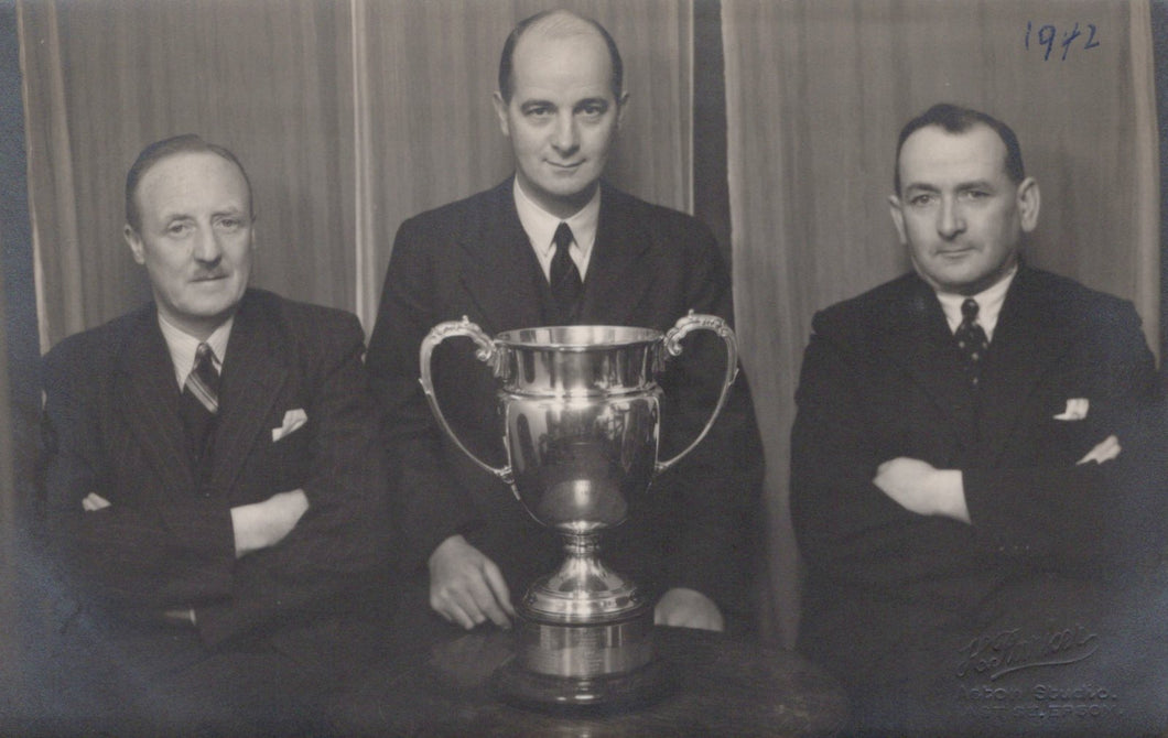 Three men in suits posing with a large trophy in front of a curtain, with '1942' written on it.