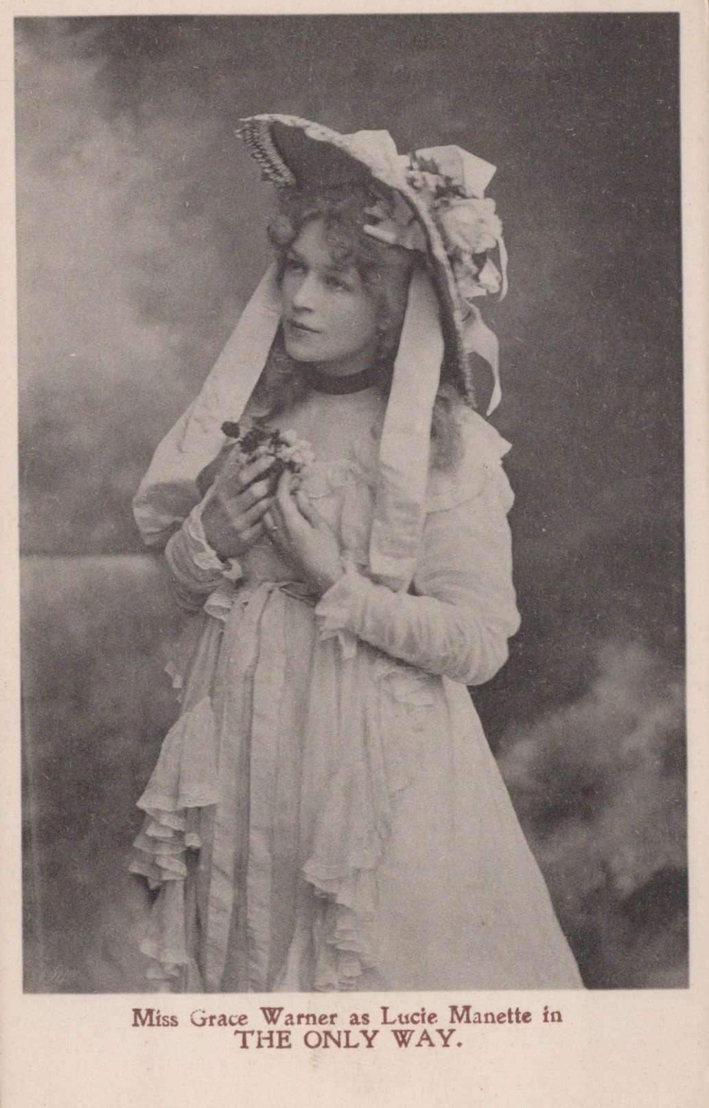 Vintage photograph of a woman in an elaborate dress and hat, holding a flower, with text at the bottom.