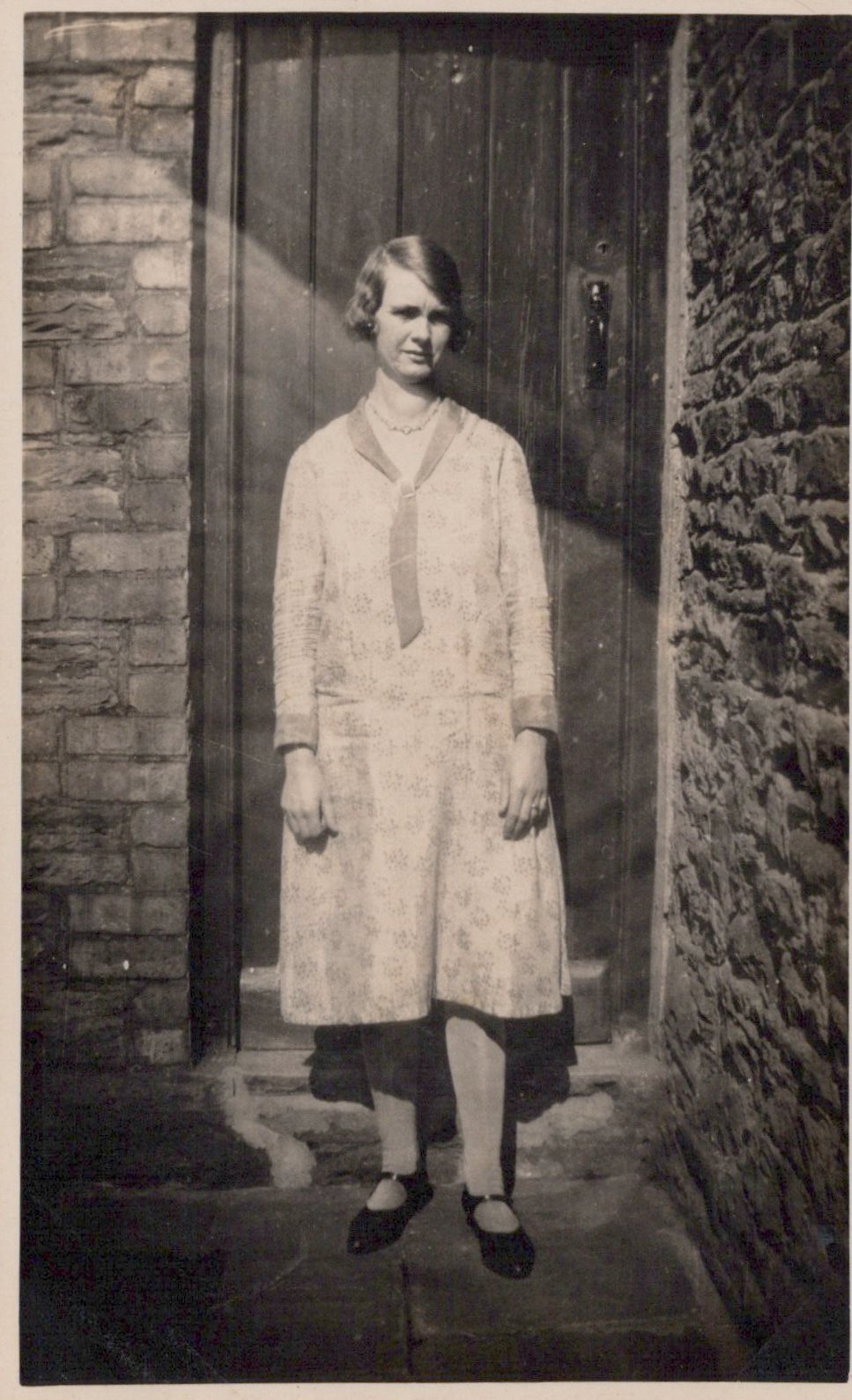 Woman in a light coat standing in front of a stone wall and door.