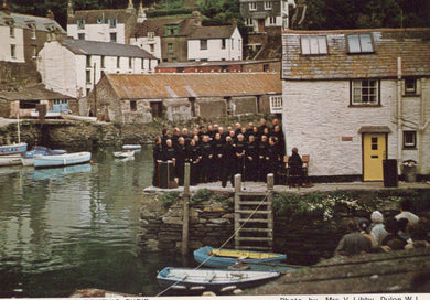 Group of people in black uniforms standing on a dock with boats and buildings in the background.