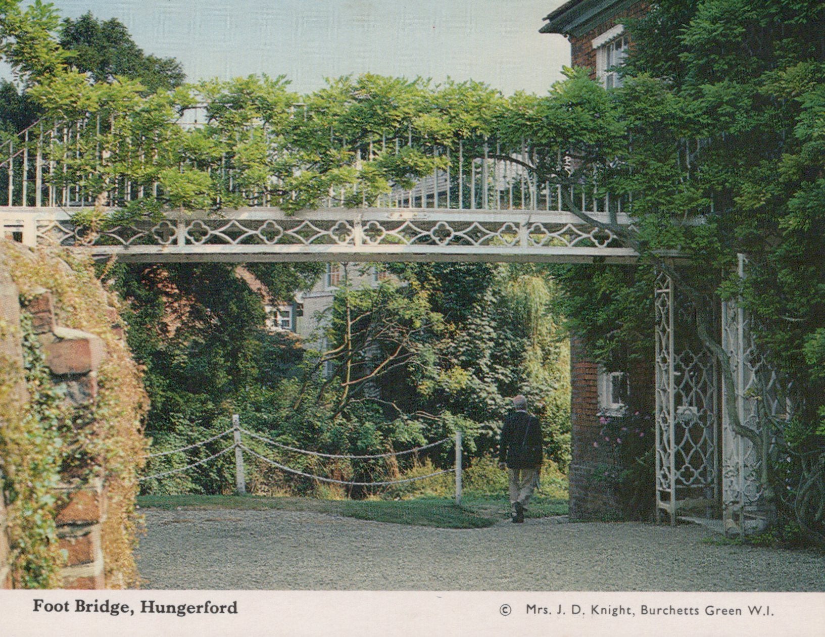 Foot bridge in a garden setting with a person walking towards it.