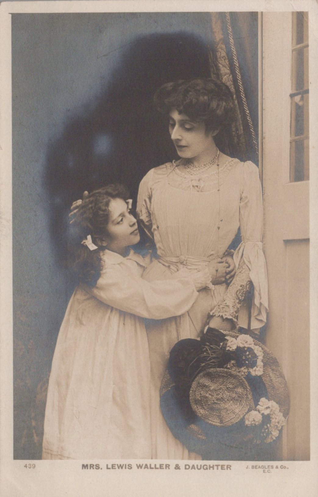 Two women in formal attire, one holding a hat, with a decorative curtain in the background.