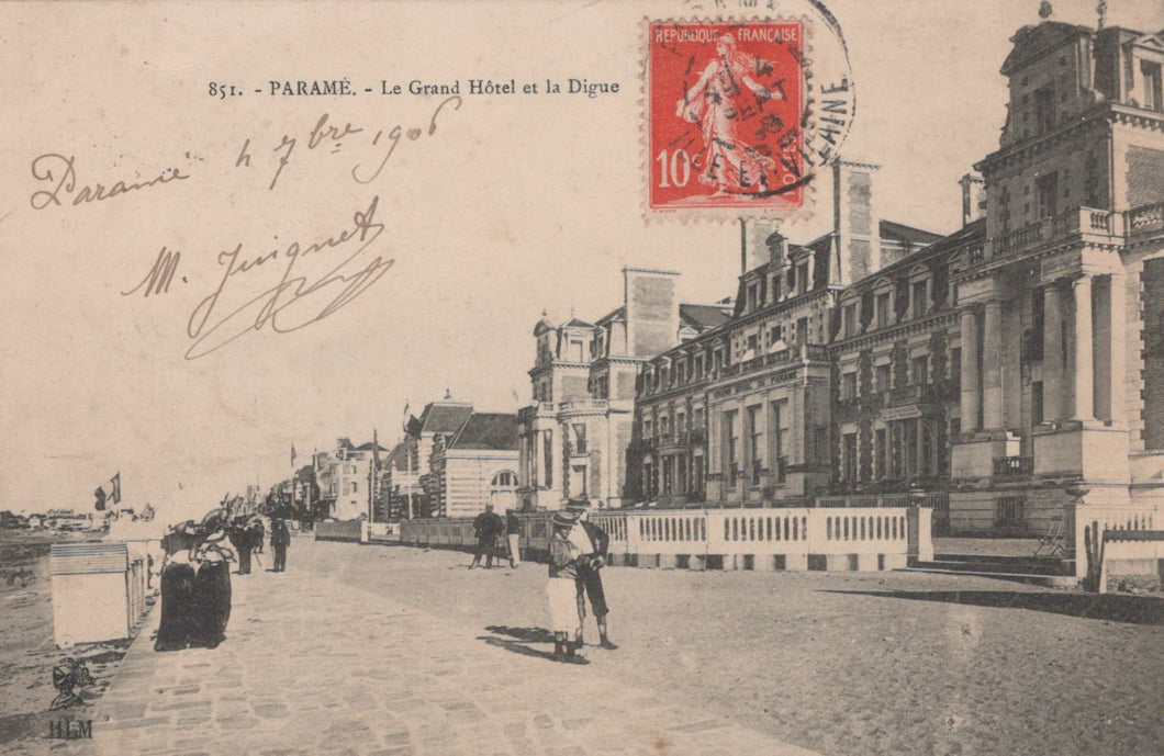 Vintage postcard of a street scene with a large building and people walking, featuring a red stamp.