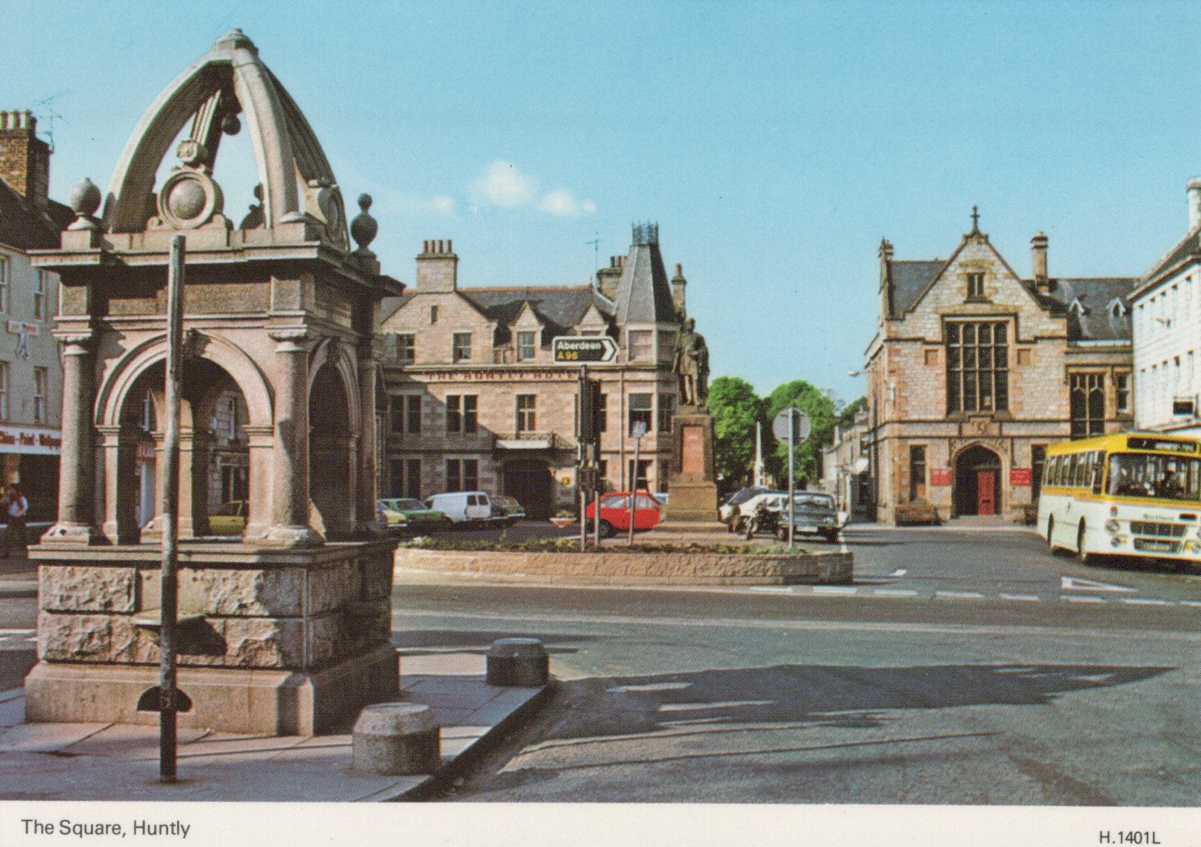 Historic stone fountain in a town square with buildings and a bus in the background.