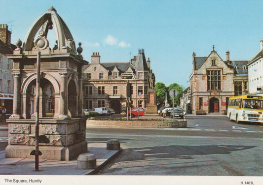 Historic stone fountain in a town square with buildings and a bus in the background.
