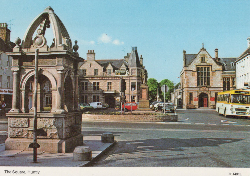 Historic stone fountain in a town square with buildings and a bus in the background.