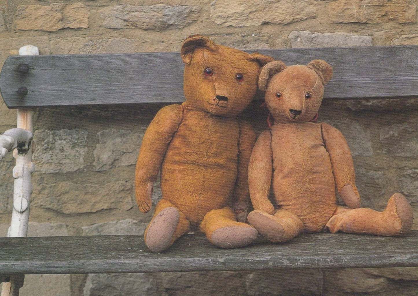 Two teddy bears sitting on a wooden bench against a stone wall.