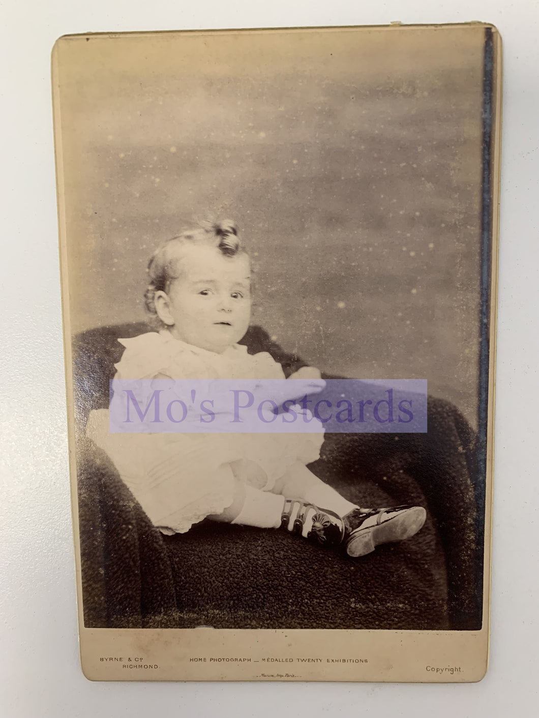 Vintage photograph of a child sitting on a chair with 'Mo's Postcards' watermark.
